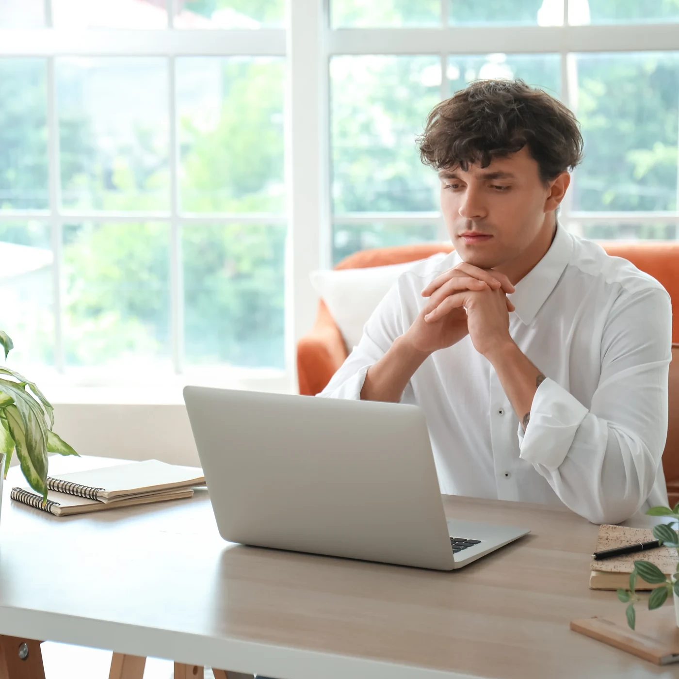 Professional thinking at a laptop beside a sunny window.